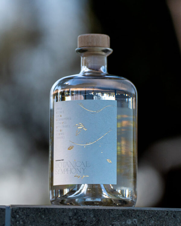 A bottle of Botanical Symphony Dry Gin standing on a stone ledge, its minimalist label with gold-foil botanical motifs glinting in soft natural light.